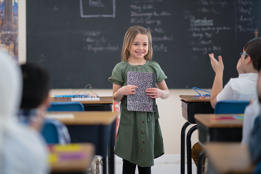 An adorable little girl of European descent smiles as she stands in front of her class presenting her work. The class is attentive and interested in what she has to say. There is some writing on the blackboard behind her.