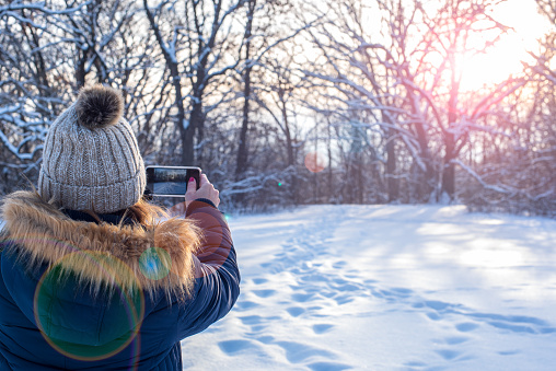 woman taking smartphone photo of sunset over winter landscape