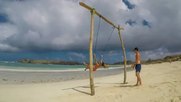 A man pushing a girl on a swing placed on the seashore of Tanjung Aan Beach, Lombok, Indonesia. The swing has simple wood construction. Waves wash the pillars of it. In the back there are few boats.