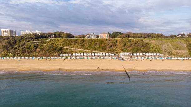 An aerial view of the Boscombe Beach with sandy beach, calm flat water, groynes (breakwaters), grassy cliff and building in the background under a cloudy sky with some blue sky An aerial view of the Boscombe Beach with sandy beach, calm flat water, groynes (breakwaters), grassy cliff and building in the background under a cloudy sky with some blue sky boscombe photos stock pictures, royalty-free photos & images