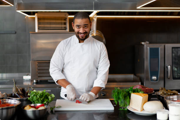 istock Handsome young African chef standing in professional kitchen in restaurant preparing a meal of meat and cheese vegetables. 1188288092