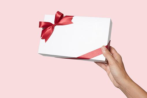 Woman hand holding holiday present white box with red ribbon on a pink background