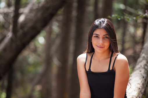 Portrait of young woman of Latin ethnicity between 18 -25 years old is in nature sitting on a tree trunk