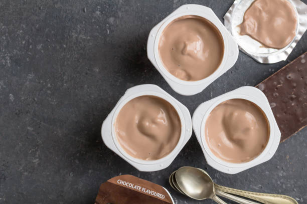 istock Chocolate yogurt cups on black mottled background with small silver spoons - Three chocolate flavoured yoghurt cups with dark chocolate bar on side - top view 1188337032