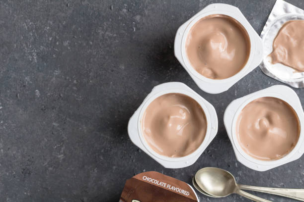 istock Chocolate yogurt cups on black mottled background with small silver spoons - Three chocolate flavoured yoghurt cups with foil lid - top view 1188339425