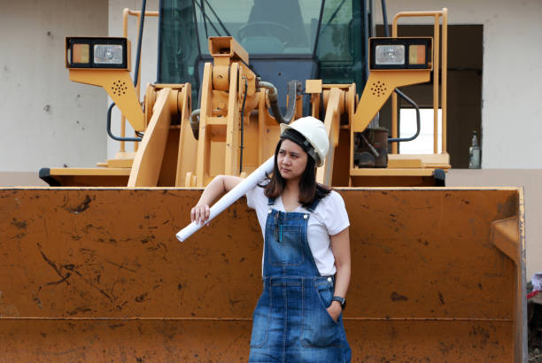 istock Female civil engineer or architect with white helmet and project drafts while in hand standing with Alloy Bulldozer Truck with Front Loader Truck Engineering Construction Car Vehicle. 1188356373