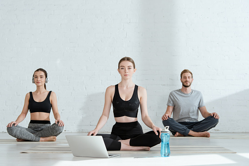 young men and woman practicing yoga in half lotus pose near laptop and sports bottle