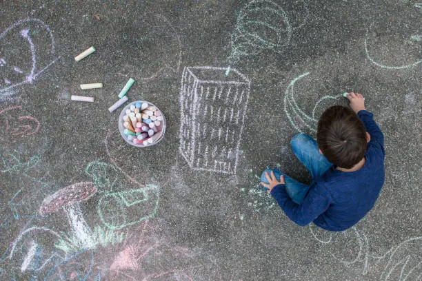 Young boy drawing outside on the sidewalk with chalk.