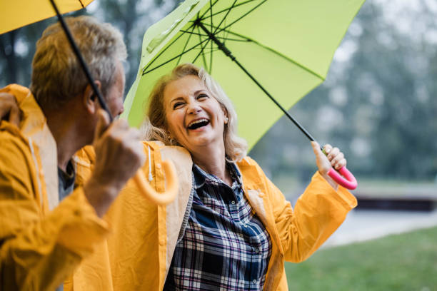 cheerful mature couple in yellow raincoats talking under umbrellas. - umbrella senior adult couple autumn imagens e fotografias de stock