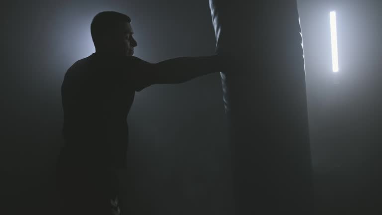 Boxer training in low light gym. Silhouette of man hitting punching bag.