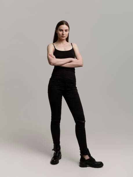 Young slim woman in black top and skinny jeans Full length budy portrait Studio shot gray background Beautiful young woman wearing black stylish clothes looking gorgeous Fashion model Full lenght body portrait on gray background arms crossed looking strong and confident sleeveless top stock pictures, royalty-free photos & images