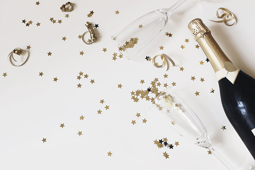 Happy New Year composition. Champagne glasses nad wine bottle with golden confetti stars isolated on white table background. Celebration, party concept, flat lay, top view. Empty copy space.