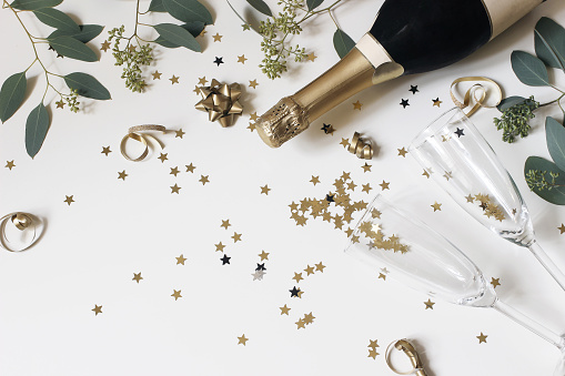 Happy New Year composition. Champagne glasses nad wine bottle with golden confetti stars and eucalyptus branches isolated on white table background, celebration, party concept. Flat lay, top view.