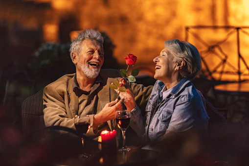 Lovely old lady admiring the red flower that her husband gave her. Couple having glass of red wine at night in roof restaurant.