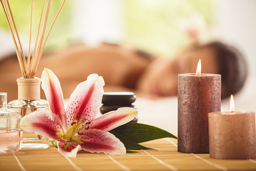 Perfumed sticks, massage oil, stones, fresh flower and scented candles on the table. Woman getting a hot stone massage in the background.