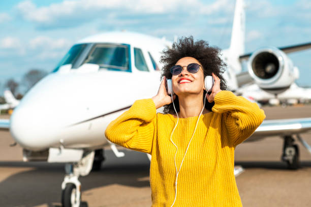 jovencita bailando alegremente frente a un avión privado estacionado en una pista del aeropuerto mientras escucha música a través de los auriculares - status symbol audio fotografías e imágenes de stock
