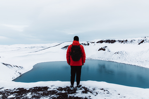Young man with backpack, in red jacket and black hat exploring the North Iceland and enjoying the view of scenic blue lake in volcano crater and snowcapped mountains