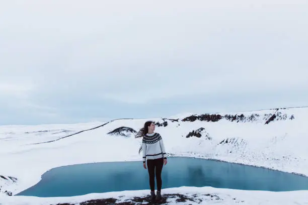 Young woman with long hair and traditional Icelandic style wool sweater exploring the North Iceland and enjoying the view of scenic blue lake in volcano crater and snowcapped mountains