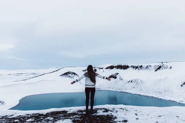 Young woman with long hair and traditional Icelandic style wool sweater exploring the North Iceland and enjoying the view of scenic blue lake in volcano crater and snowcapped mountains