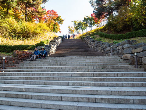 Seoul, South Korea - November 4, 2019: couple of elderly tourists rest on the steps of the stairs and a group of guys descend from above in colorful city park in Seoul city in sunny autumn morning