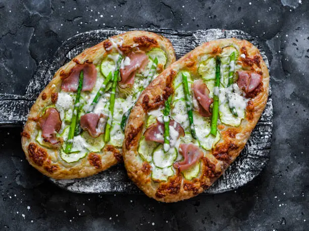Photo of Puff pastry ham, zucchini, asparagus, mozzarella cheese pizza no wooden rustic chopping board on dark background, top view