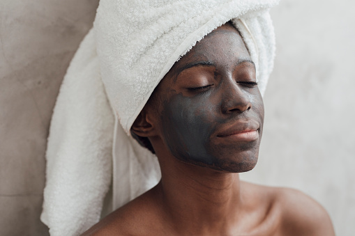 Portrait of pretty relaxed African woman wrapped in bath towels with black face mask.