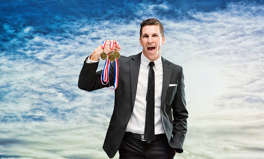 One man only / one person / waist up / front view of 30-39 years old handsome people caucasian male / young men businessman / business person / manager standing in front of nature / scenics - nature who is outdoors wearing businesswear / a suit who is smiling / happy / cheerful and showing award who is in first place and holding medallion / gold medal / medal / gold / sky