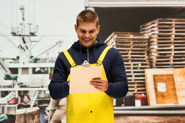Waist up / one man only / one person of 20-29 years old adult handsome people caucasian male / young men standing who by the sea wearing hooded jacket / shirt / sweatshirt / bib overalls who is working and holding clipboard / document / paperwork / contract / to do list / nautical vessel cintura para arriba / un hombre solamente / una persona de 20-29 años de edad adulto guapo personas caucásicas hombres / jóvenes de pie que por el mar usando chaqueta con capucha / camisa / sudadera / babero general que está trabajando / barco náutico