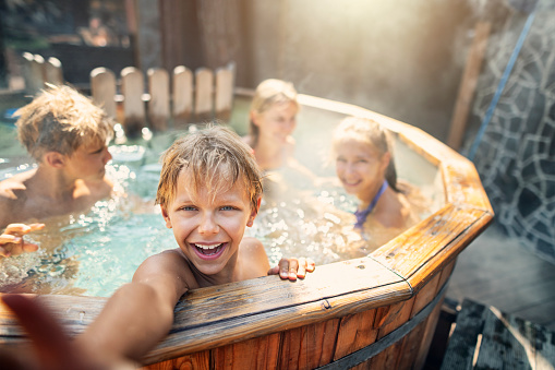 Family is playing in wood fired barrel hot tub in the back yard. Boy is laughing at reaching at the camera.\nNikon D850