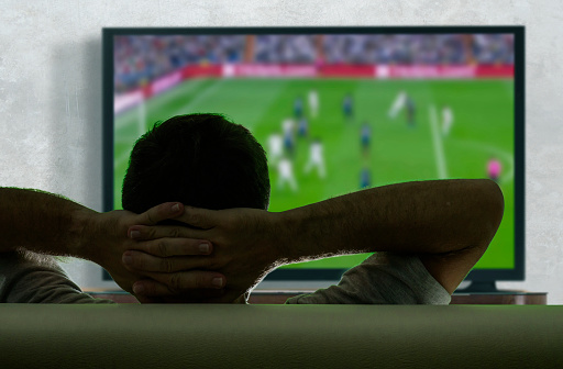 view from behind of young and happy soccer fan man watching European football game on big screen television at living room sofa couch enjoying and feeling the emotion cheering his team