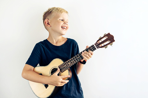 Young blond teenager plays ukulele and sings a fun song. Portrait of a laughing child engaged in a favorite hobby on a big background with place for text.