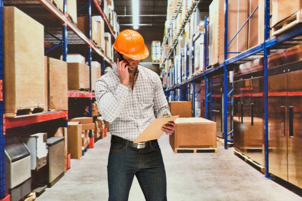 One man only / one person / waist up / front view of male / young men manual worker / photography standing at the warehouse / distribution warehouse / factory / storage compartment / storage room in front of merchandise wearing helmet / work helmet / hardhat who is talking who is delivering and holding box - container / package / cardboard / cardboard box / container / clipboard / contract / mobile phone / using smart phone / packaging / pallet / shipping un hombre sólo / una persona / cintura para arriba / vista frontal de hombres / hombres jóvenes trabajador manual / fotografía de pie en el almacén / almacén de distribución / fábrica / compartimento de almacenamiento / trastero frente a la mercanc�