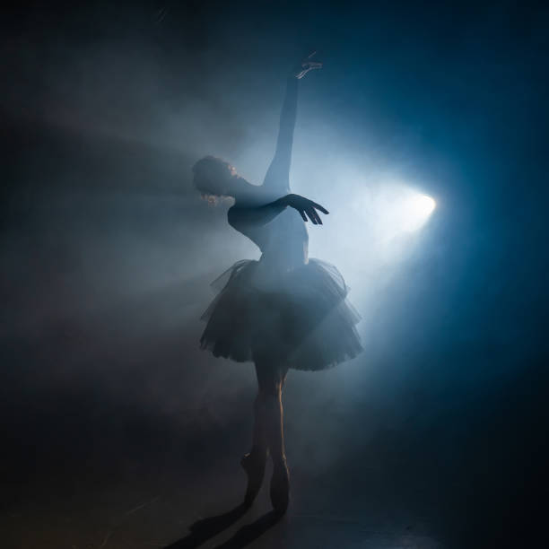 istock Young beautiful ballerina on smoke stage dancing modern ballet. Performs smooth movements with hands against spotlight background. Woman in black tutu costume on scene. 1188460655