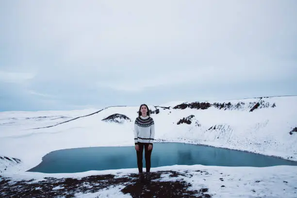 Young woman with long hair and traditional Icelandic style wool sweater exploring the North Iceland and enjoying the view of scenic blue lake in volcano crater and snowcapped mountains