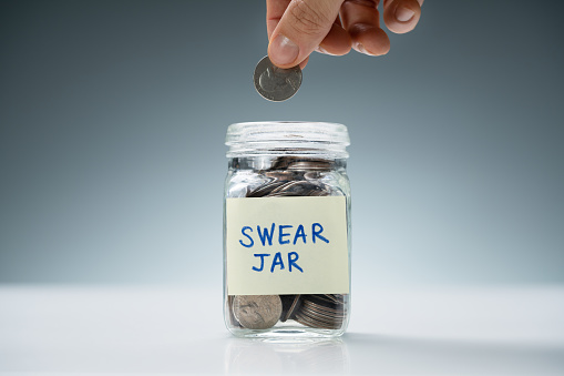 Person's Hand Inserting Coin In Glass Jar With Swear Jar Text Over White Desk