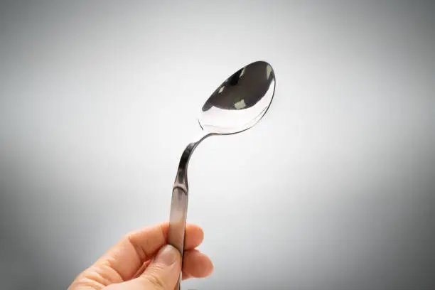 Close-up Of A Person's Hand Holding Bend Stainless Steel Spoon Against Gray Background