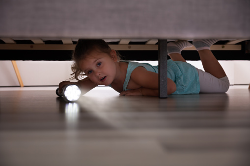 Little Girl Lying Under The Bed Searching For Something With Flash Light At Home