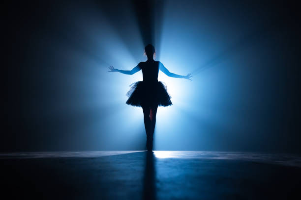 istock Young beautiful ballerina on smoke stage dancing modern ballet. Performs smooth movements with hands against spotlight background. Woman in black tutu costume on scene. 1188464814