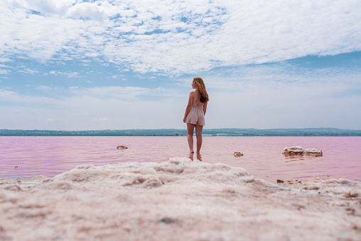 Back view of cute teenager woman wearing summer clothes standing on an amazing pink lake of sea water and blue sky