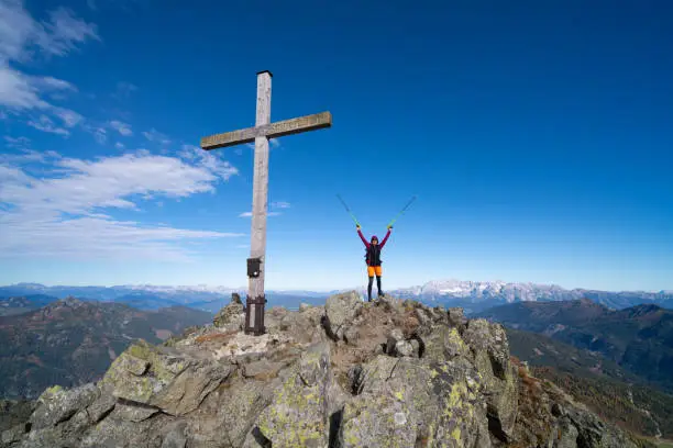 happy mature mountaineer hiking cross country running woman standing alone high up in beautiful alpine mountain landscape on mountain peak near summit cross with arms and trekking poles raised up