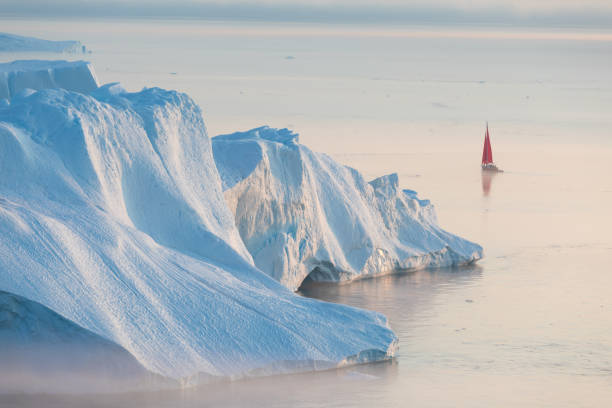Little red sailboat cruising among floating icebergs in Disko Bay glacier during midnight sun season of polar summer. Ilulissat, Greenland. Unesco world heritage Disko Bay during midnight sun Ilulissat, Greenland. Studying of a phenomenon of global warming Ices and icebergs disko bay stock pictures, royalty-free photos & images