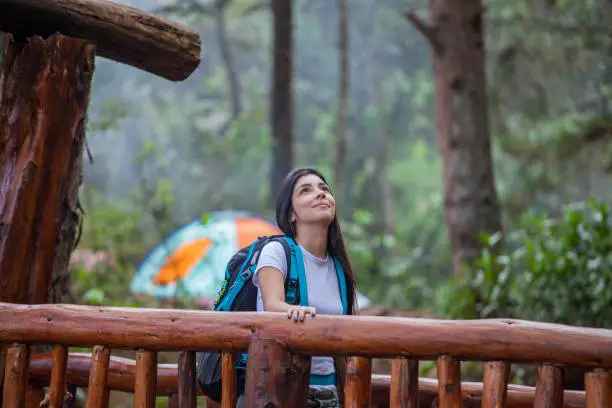 Young latin woman with long black hair in white blouse and travel backpack walks over wooden bridge located in the natural park of the city of Medellin
