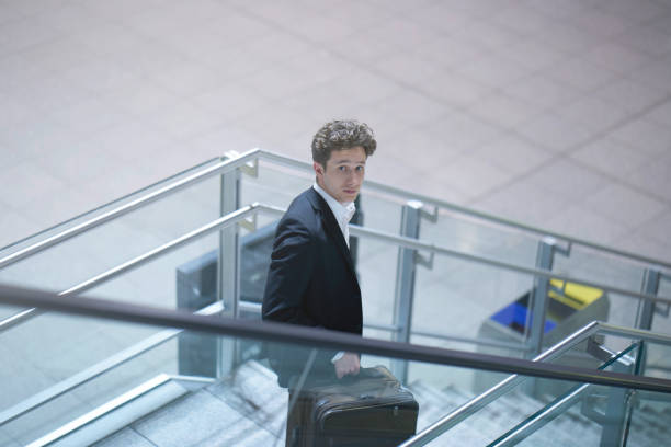 istock Young businessman going down a stairway with his suitcase, looking up, on the move, wearing a business suit 1188481767