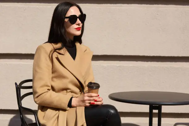 Brunette attractive young female sitting at table outdoors, looking aside, wearing beige coat, black trousers and sunglasses, holding papercup of coffee in hands, having lunchtime. Relax concept.