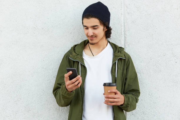 Portrait of peaceful slender youngster posing isolated over white wall background outdoors, surfing net, checking social networking sites, holding smartphone and papercup of hot drink in hands. retrato de joven restito pacífico posando aislado sobre fondo de pared blanca al aire libre, la red de surf, la retención de sitios de redes sociales, la celebración de teléfonos inteligentes y papel cupo de bebida caliente en las manos.