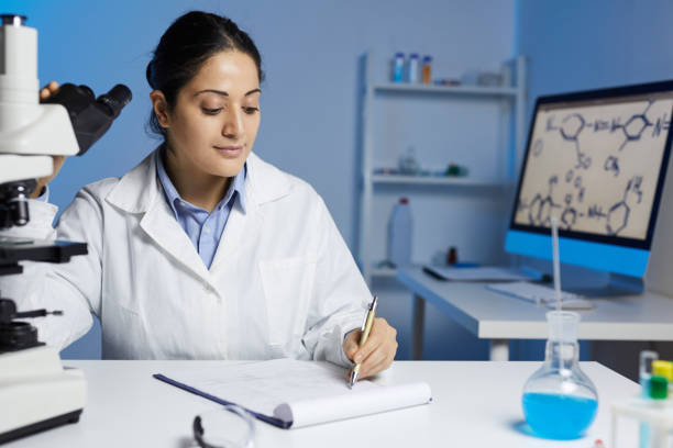 istock Content young Indian female scientist in lab coat sitting at table and making notes on research 1188486054