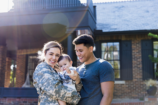 Happy mid adult Hispanic female soldier smiles at the camera during a sweet family reunion. They are standing outdoors in front of their home.