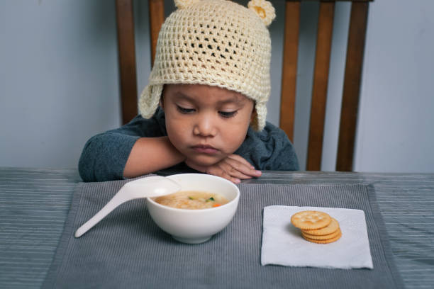 Loss of appetite for a sick toddler who is holding his sore throat with his hand and is staring at a bowl of homemade soup. stock photo