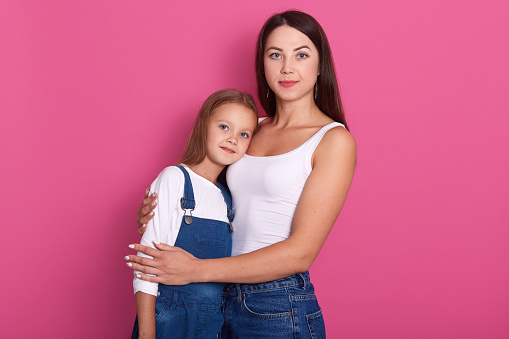 Studio portrait of attaractive young mother hugging her small daughter, females wearing white shirts and jeans, posing isolated over pink studio background, looking at camera. Family concept.