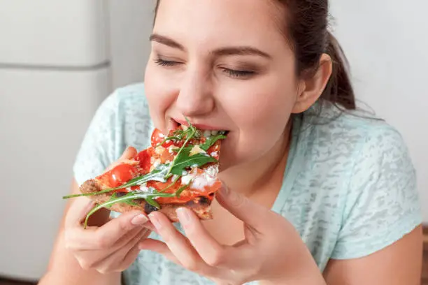 Young chubby woman sitting at table in kitchen binge eating eating slice of pizza close-up closed eyes smiling delightful